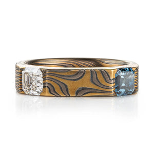 unique mokume gane band with flat top and angled corner settings, the stones are both square asscher cuts, diamond and blue sapphire, ring is made with yellow gold, palladium and silver