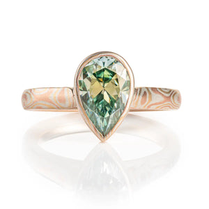 mokume gane ring with green moissanite in a pear shape, the stone is held in a mokume bezel, bezel and ring are patterned in woodgrain, with red gold yellow gold and silver