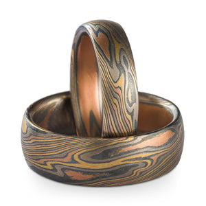 A set of Mokume Gane rings or wedding bands made by Arn Krebs, one is laid down flat on the surface, the other is propped up perpendicular inside the first. The rings are made in our twist pattern and firestorm palette, this palette is palladium silver red gold and yellow gold, both rings have low dome profiles