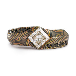 This is a custom made Mokume Gane ring, made by arn krebs, featuring a unique carved angled profile and a modified cathedral style setting holding the large white center diamond. The diamond is square and set diagonally to the band, and is flanked on each side by seven small bead set black diamonds running parallel to the band. It's made in our Twist pattern and an upgraded 18kt version of our Firestorm palette, the metals in it are red gold yellow gold palladium and silver