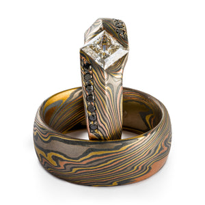 Set of rings, Mokume Gane, custom made, ring laying flat is a plain band, ring propped upright is a custom cathedral style setting with a large white center diamond flanked by rows of black diamonds bead set, both rings are firestorm palette and twist pattern