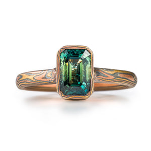 magical feeling woodgrain patterned narrow ring with a single green rectangular stone, held in a mokume patterned bezel - a narrow strip of the same material as the main ring wrapped around the perimeter of the stone