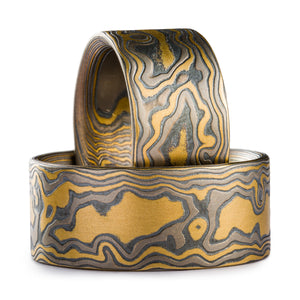 Set of Mokume Gane rings made by arn krebs, one ring is propped up inside the other one which is laid flat. Both are made in a woodgrain pattern and are made of layers of yellow gold palladium and oxidized silver