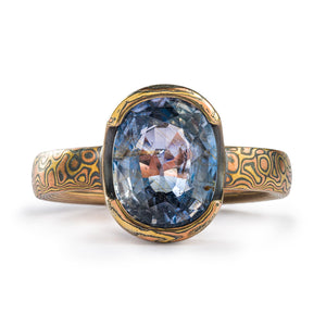 Mokume gane engagement or wedding ring, droplet pattern, large oval shaped blue sapphire held in a partial bezel. Everything including the bezel is mokume gane, with a droplet style pattern. Metals used in the ring are red gold yellow gold and oxidized silver