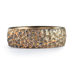 Mokume gane style ring, 7mm wide, pattern runs diagonally across the band with multicolored metal layers, center of pattern is droplet/spots, sides are more straight lines, and whole ring has carved surface texture meant to imitate hammering