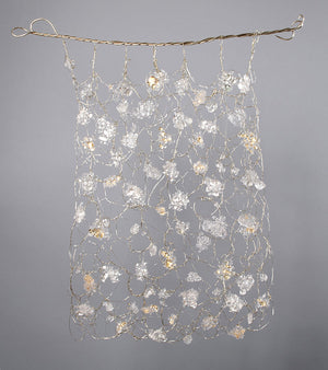 Hand woven wire tapestry with many cast glass pieces interwoven, with small gold accents throughout. Tapestry hangs from a twined metal bar, overall white and gold color palette. Elegant home decor, fine arts, home art collection, interior design.