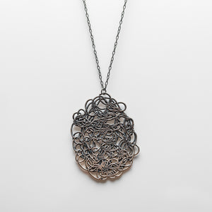 Pendant made of woven wire spun into an oval shape, pressed flat. Darker gray towards the top, turning lighter at the bottom. Hanging on a silver chain.