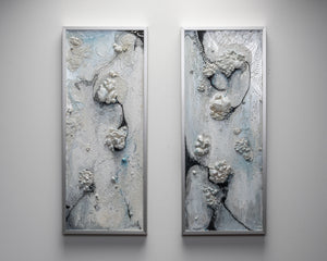 Pair of framed rectangular art pieces hanging on a white wall. Both pieces are mixed media, with an icy blue base with black accents and three dimensional pieces that resembles bubbles.