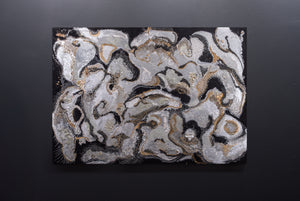 Large rectangular artwork hung on a gray wall, made of swirling black, silver and gold, with textured details.