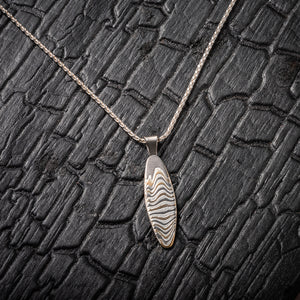 Slim oval shaped mokume gane patterned pendant, made with layers of yellow gold, palladium and silver, on a silver chain. 