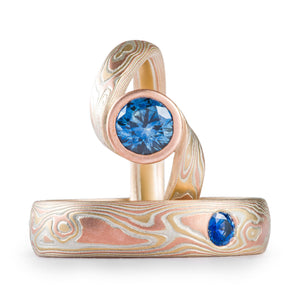 Set of rings with blue sapphire stones, both in mokume gane metal combination of red gold yellow gold and silver with a woodgrain pattern, ring with larger stone is set in a bypass style with twisted shank