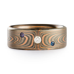 Mokume Gane 8mm wide ring made by arn krebs, vortex pattern and embers palette, flat profile, with flush set stones. The embers palette is made of red gold palladium and silver and the ring has been etched and oxidized. The stones set are a central and larger diamond, and on either side are a tanzanite and sapphire, both blue, so there are 3 stones total in the ring. 
