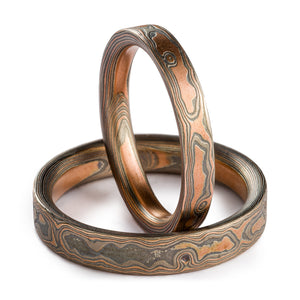 Mokume Gane ring set, woodgrain pattern and embers palette. Embers palette is red gold palladium and silver, both rings are etched and oxidized and have a flat profile. One ring is laid flat on the surface, second ring is propped up vertically in it. 