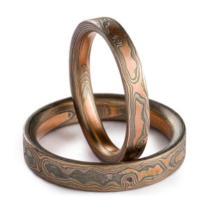 Mokume Gane ring set, woodgrain pattern and embers palette. Embers palette is red gold palladium and silver, both rings are etched and oxidized and have a flat profile. One ring is laid flat on the surface, second ring is propped up vertically in it. 