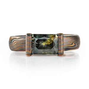 whimsical and unique engagement style mokume gane ring, shown in Firestorm palette with matching mokume patterned bezel, holding a rectangular mossy green sapphire