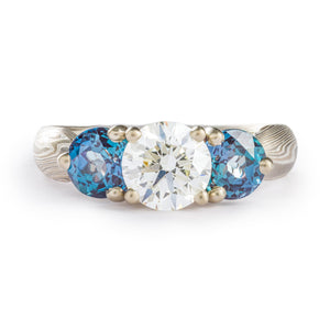 Emgagement style ring, 3 head prong setting in the center, a slightly larger round white diamond in the middle, two smaller blue gems on either side (alexandrites), prongs are white gold, the ring is an overall silver color, the mokume is made of silver palladium and white gold