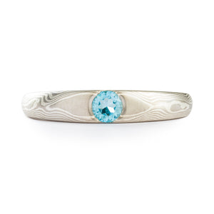 Ring with single light blue circle shaped stone in the center, 4mm at center and tapers gently from that point. Mokume Gane style cathedral ring by Arn Krebs, pattern resembles the grain of wood, and is made up of white gold, palladium and silver. 