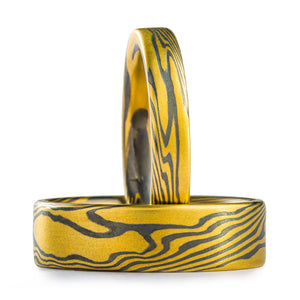 Two matching mokume gane twist pattern rings, one laid flat the other upright, both are made with yellow gold and oxidized silver and have flat profiles. The laid down ring is wider than the upright one.
