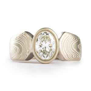 Unique modified cathedral style ring with mokume gane patterning, the main band is split in the center with gently curved ends, and is holding an oval bezel with a diamond that is the same height as the band. The ring is overall silver and white
