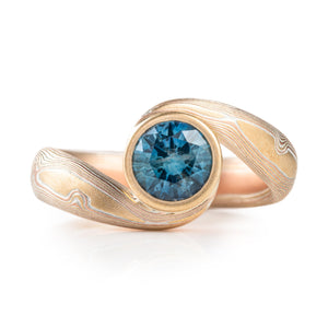 deep blue sapphire set in a bypass setting where the mokume is swirled around the stone and holds it in Place in a swooping circle . the mokume is red gold and yellow gold and silver
