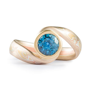 teal blue sapphire set in volume woodgrain that is a yellow gold and red gold palette . handmade goldsmith made ring