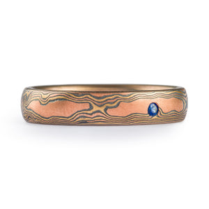 mokume gane band in woodgrain pattern, red gold yellow gold and oxidized silver with a small flush set blue sapphire