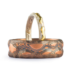 Two rings, one larger and wider that's mainly red gold with oxidized silver, the smaller and thinner band is palladium and silver with a yellow gold raised area in the center line of the band.