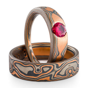 Mokume Gane ring set or wedding ring set made by arn krebs, both rings have flat profiles and are made in the woodgrain pattern and embers palette. The plain band is laid down and the engagement style ring is stood up perpendicular to the other ring and has a cathedral style set ruby. The embers palette is made of red gold, palladium and silver, and the plain band has an added palladium top sheet. 